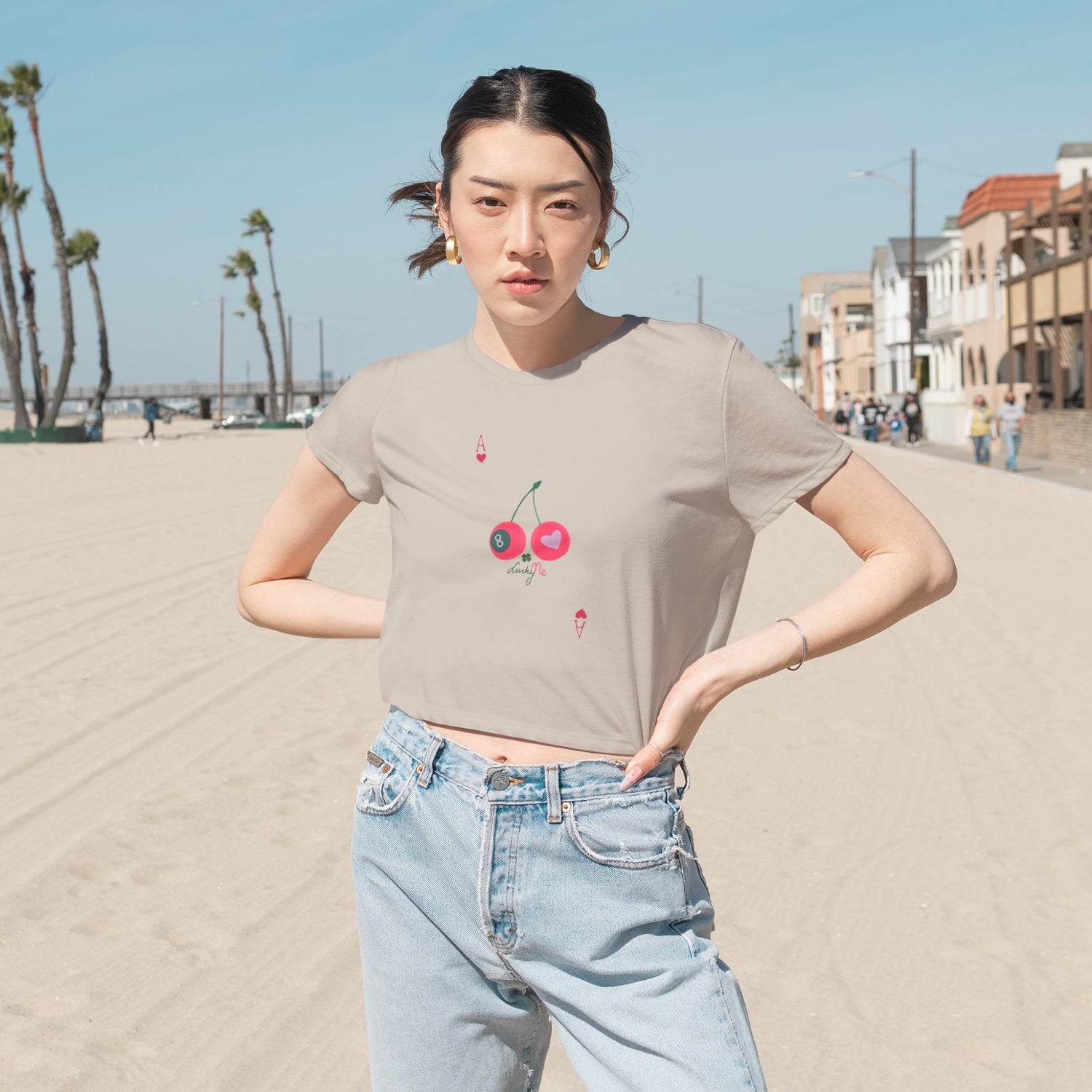 Woman wearing the heather dust Lucky Me cropped tee with cherry and 8-ball print, styled with jeans at the beach.