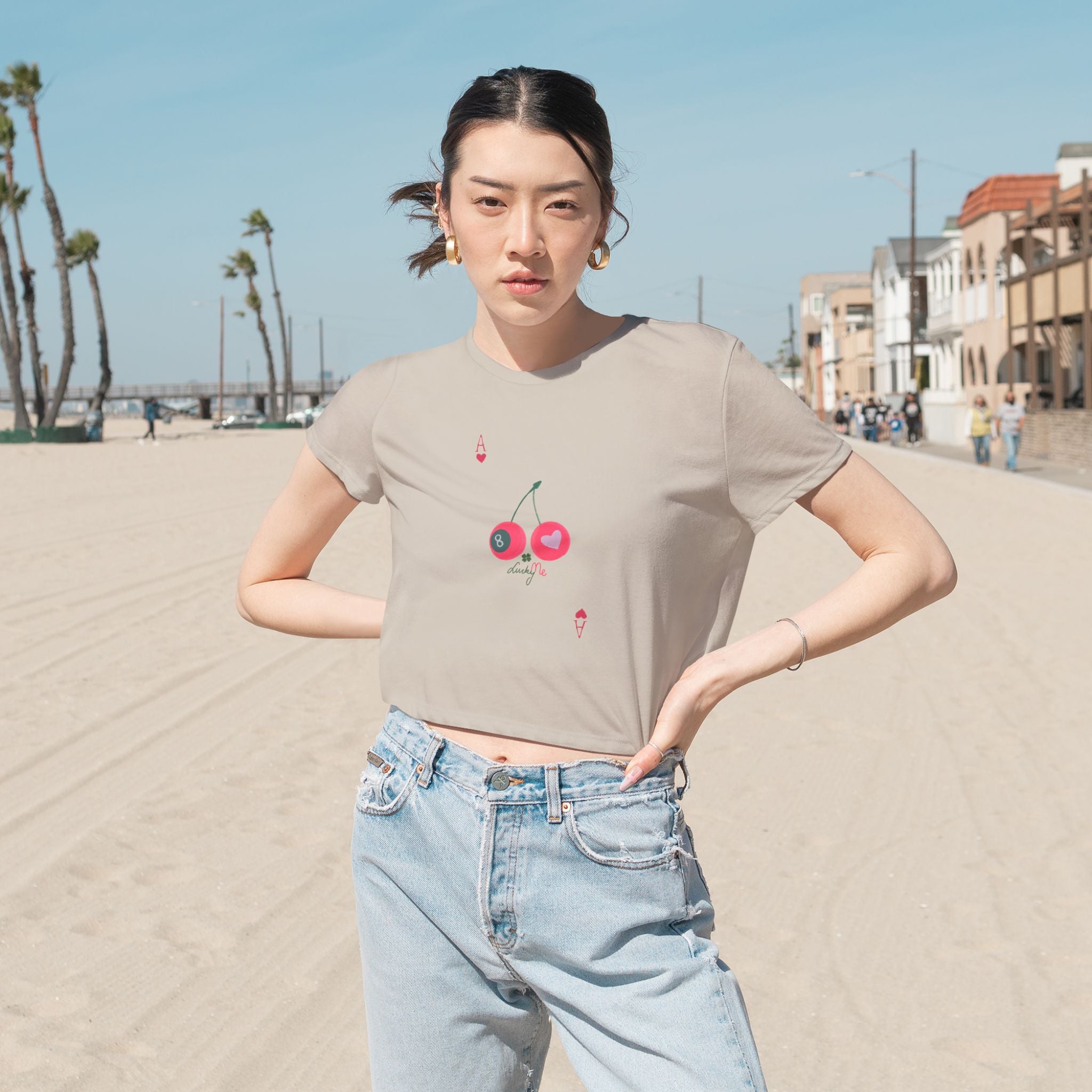 Woman wearing the heather dust Lucky Me cropped tee with cherry and 8-ball print, styled with jeans at the beach.
